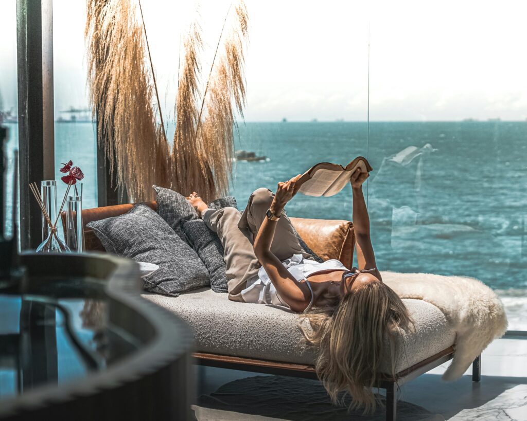 A woman reading while lounging on a couch next to a glass wall with a stunning ocean view.